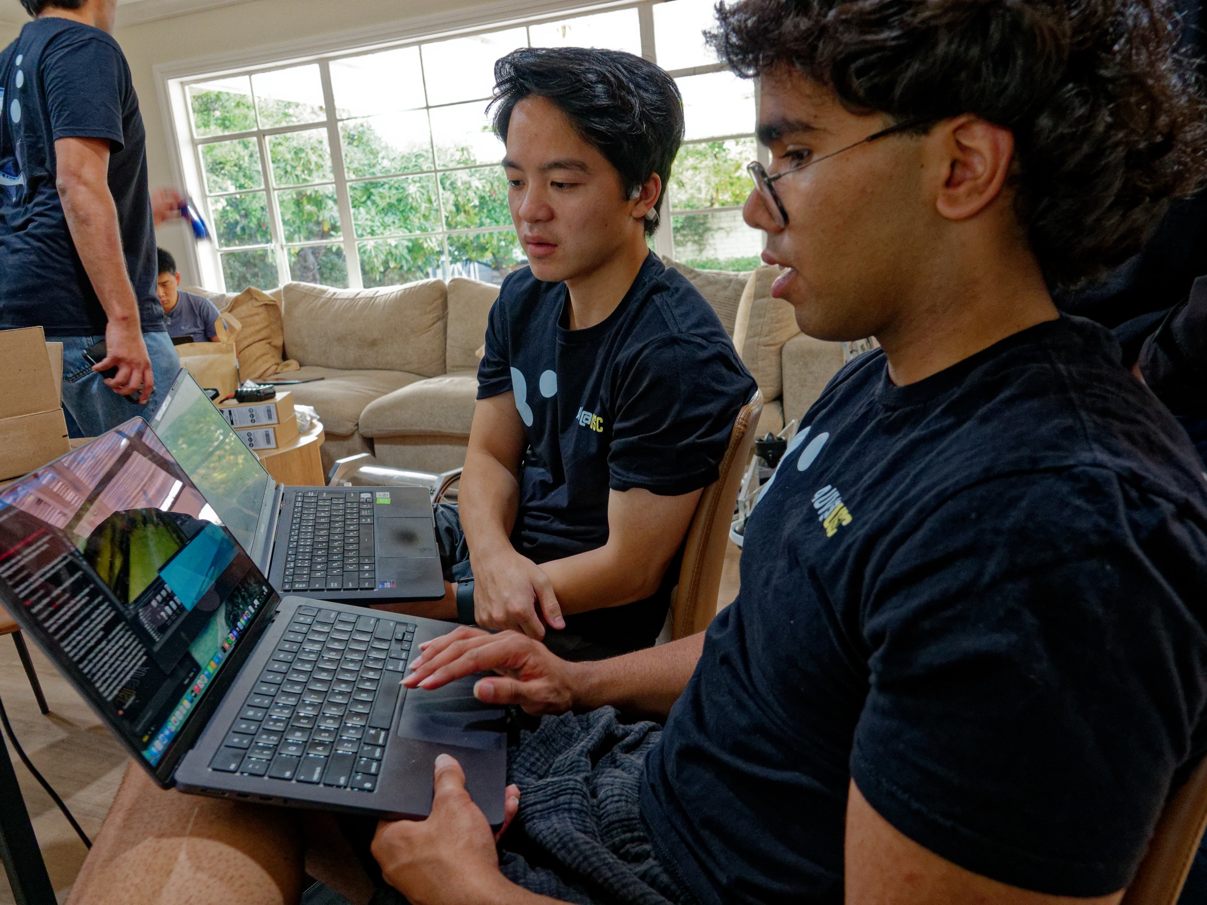 Team members reviewing camera feeds and code on their laptops while preparing at the team house.